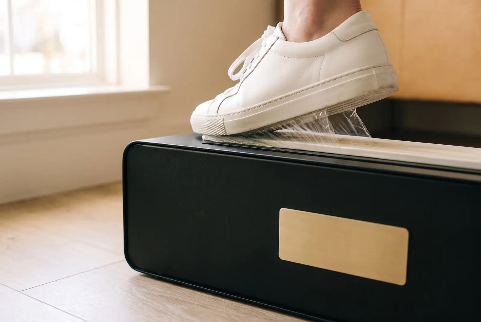 A shoe pressing down on the plate of a TidySteps unit at the door, the clear film held open beneath it.